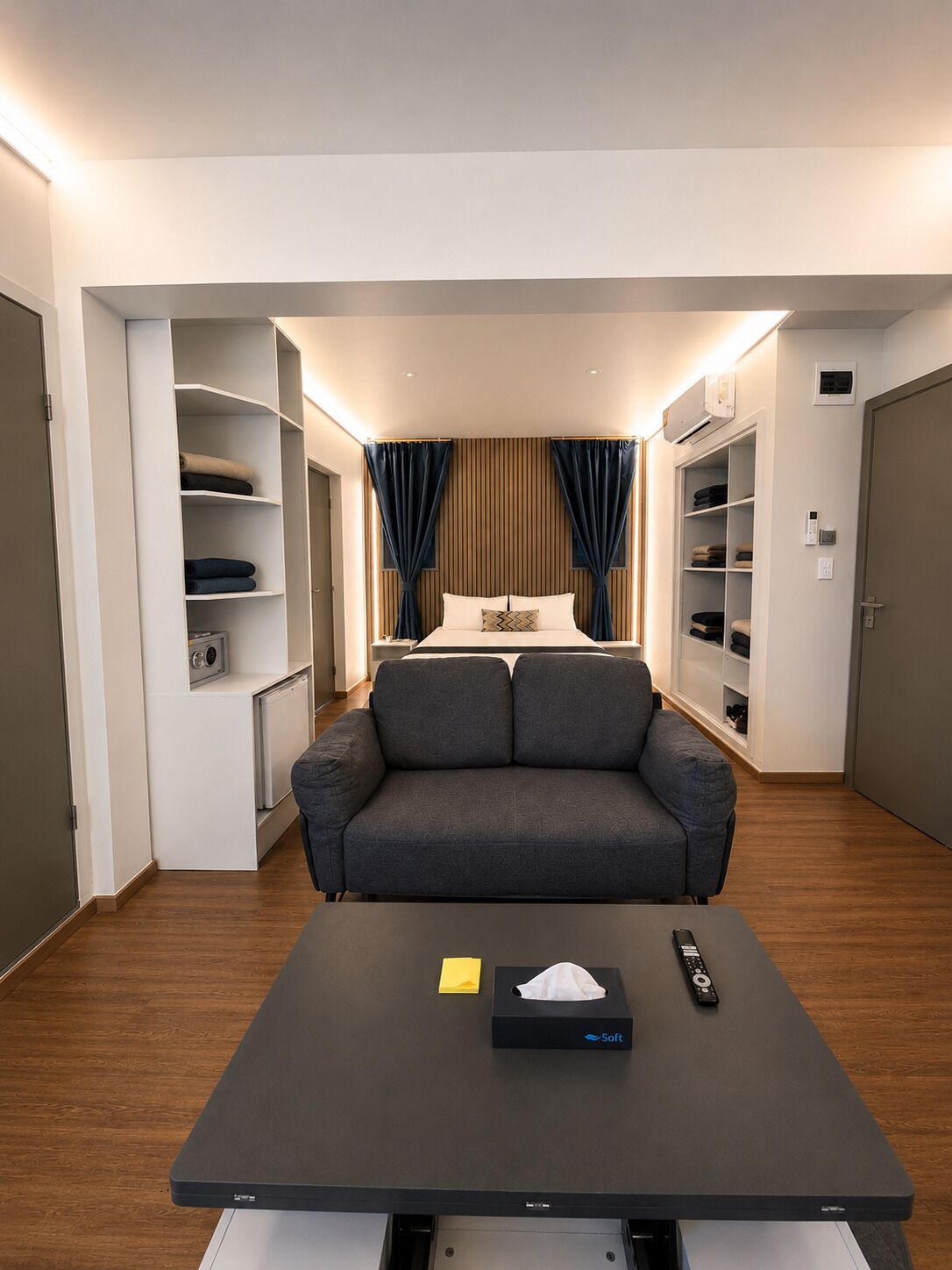 Habitación de hotel moderna con sofá, una mesita de noche con control remoto, caja de pañuelos y almacenamiento empotrado.