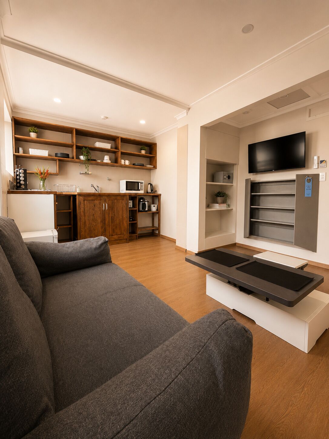 A modern living room with a sofa, a flat-screen TV, and a kitchen area with wooden shelves and appliances.