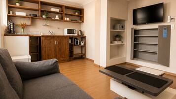 A modern living room with a sofa, a flat-screen TV, and a kitchen area with wooden shelves and appliances.
