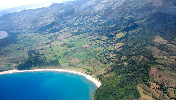 Vista aérea de uma paisagem costeira com praia, colinas ondulantes e vegetação dispersa.
