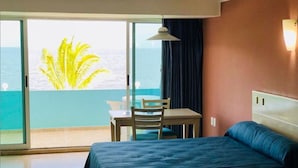 Camera d'albergo con un letto, un tavolo e una sedia. Si vede l'oceano attraverso un'ampia finestra.