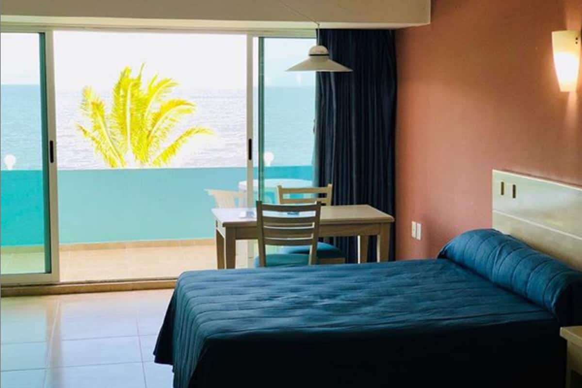 A hotel room with a bed, a table, and a chair. There is a view of the ocean through a large window.