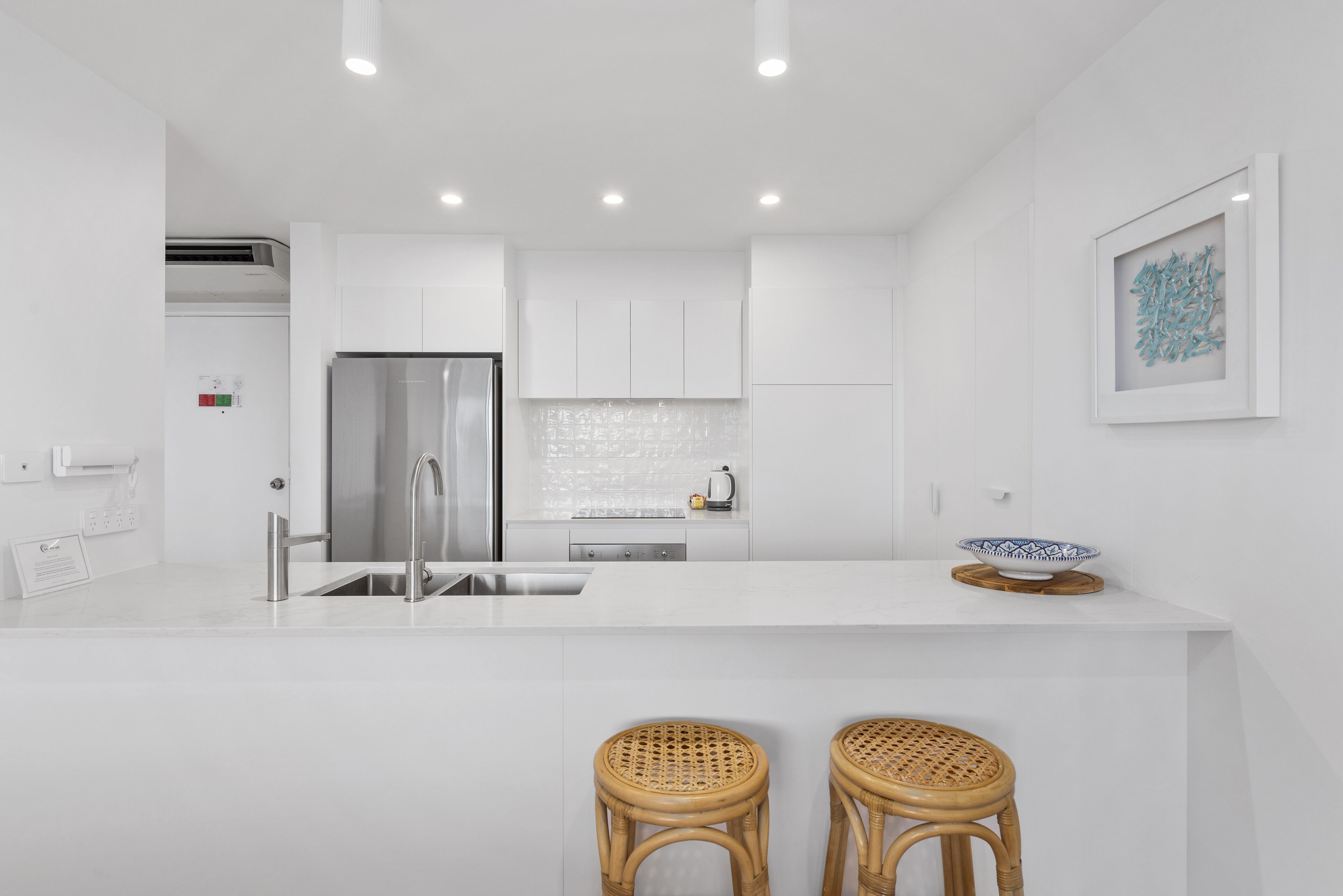 A modern kitchen with white cabinetry, a stainless steel refrigerator, a sink, and two wicker stools.