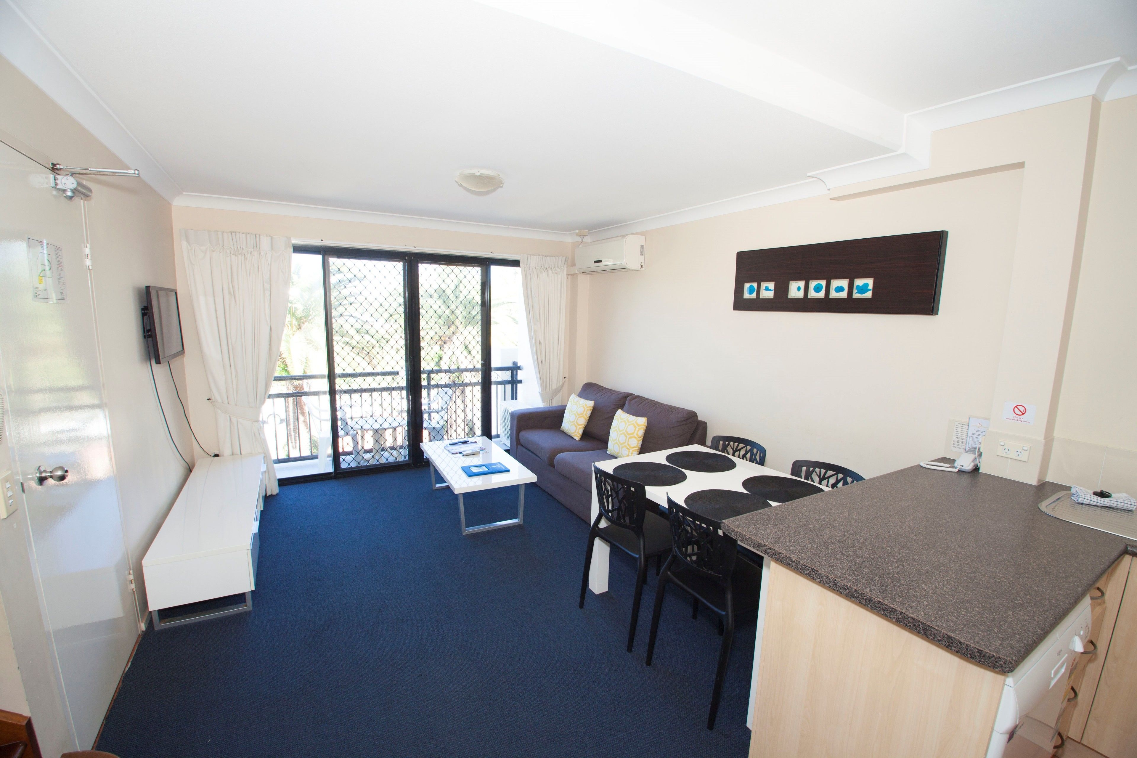 A compact living space with a sofa, dining table, chairs, a small kitchen area, and a sliding glass door leading to a balcony.