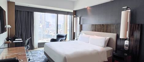 A hotel room with a large bed, a desk with a chair, a desk lamp, and a view of the cityscape.