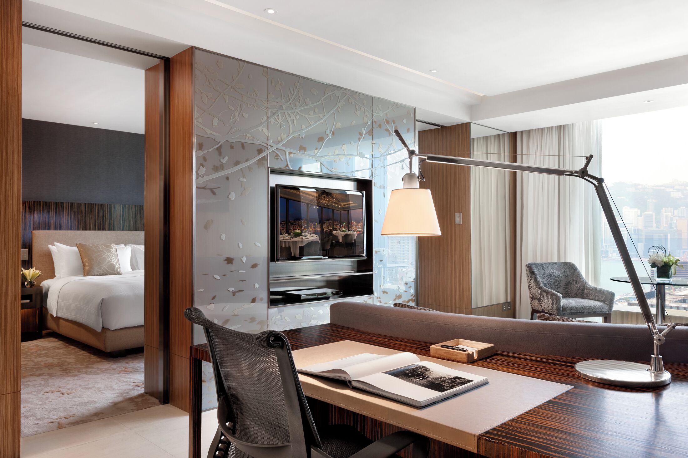 A modern hotel room with a bed, a seating area with a table, a TV, and a city view.