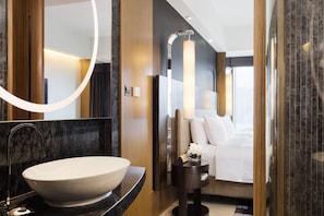 A modern bathroom with a large oval sink, a mirror, and a bedside table with a lamp.