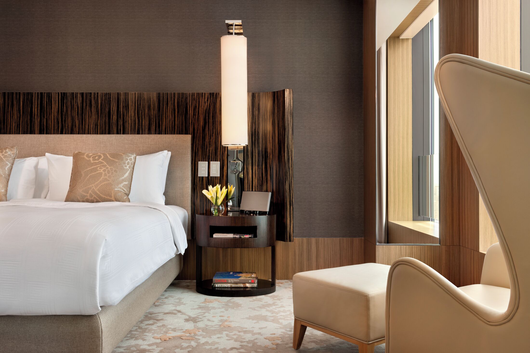 A modern hotel room with a large bed, a bedside table with a lamp, a chair, and a view of the outdoors.