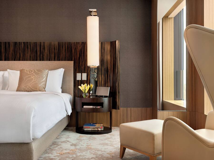 Quarto de hotel moderno com uma cama grande, uma mesa de cabeceira com abajur, uma cadeira e vista para o exterior.