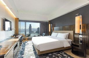 A hotel room with a large bed, a desk, a chair, a lamp, and a view of the cityscape.
