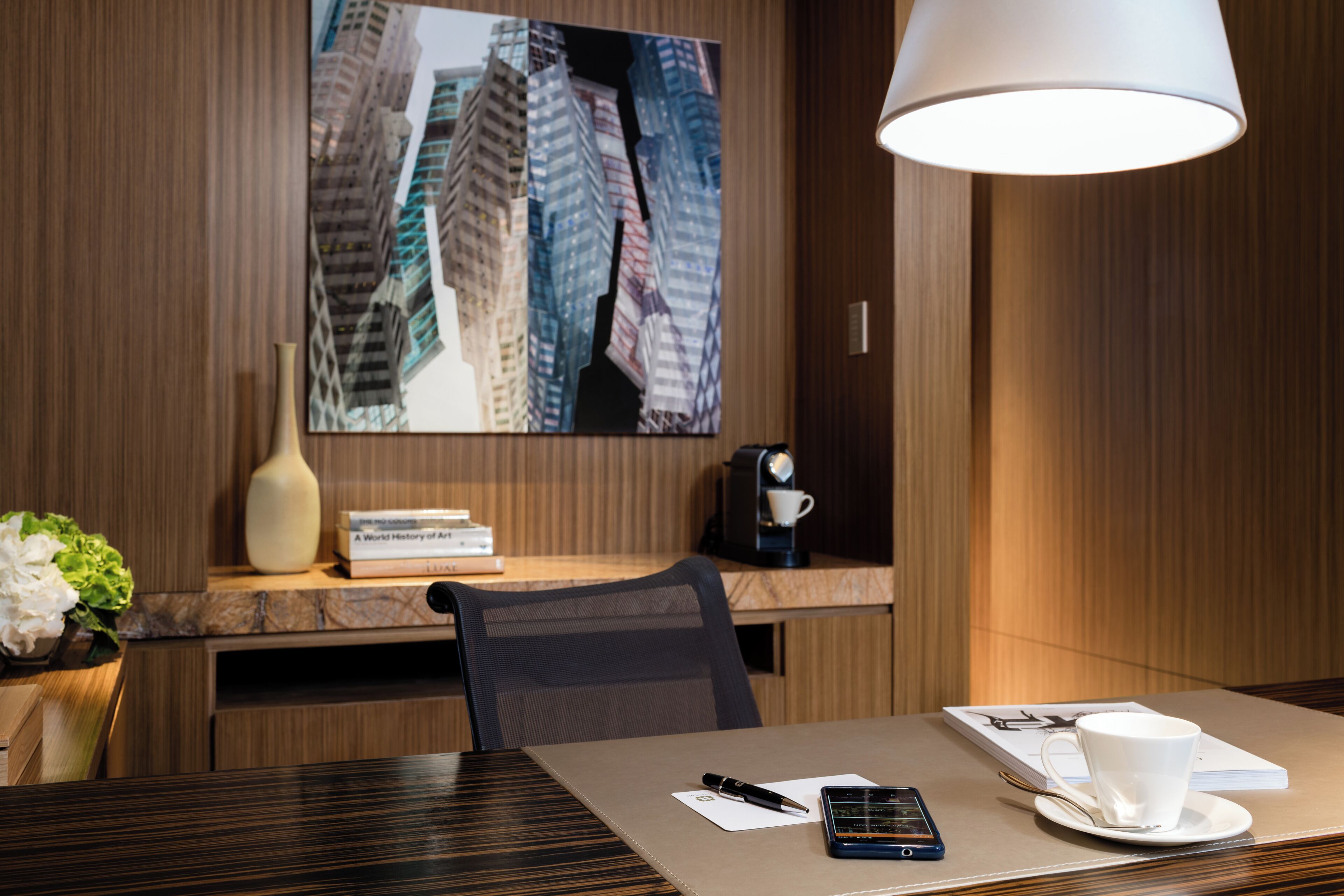 A hotel room with a wooden desk, a coffee machine, a white cup, a smartphone, and a book titled 'A World History of Art'.