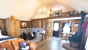 A rustic room with a bed, a wooden dining table, a chandelier, and a kitchen area with a stove.