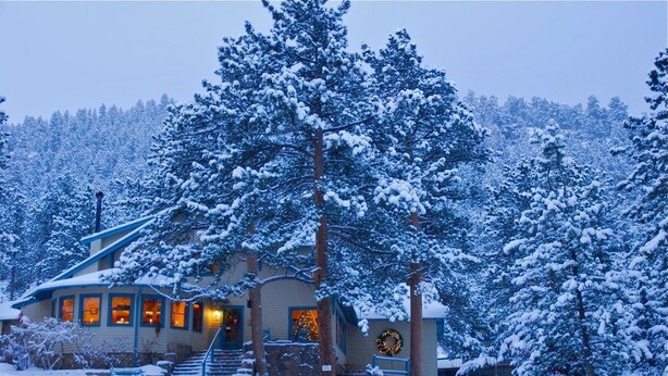A snow-covered lodge with a staircase leading to the entrance, surrounded by tall trees.