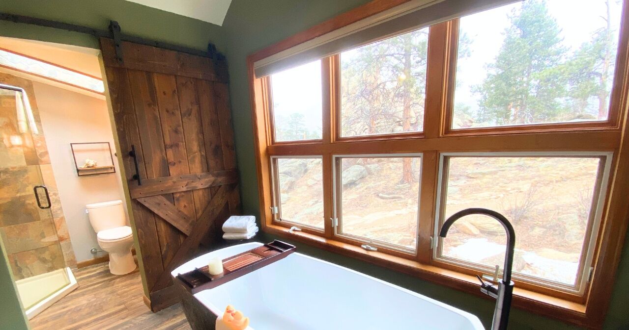 A bathroom with a bathtub, a window with a view of trees, a wooden door, and a toilet.