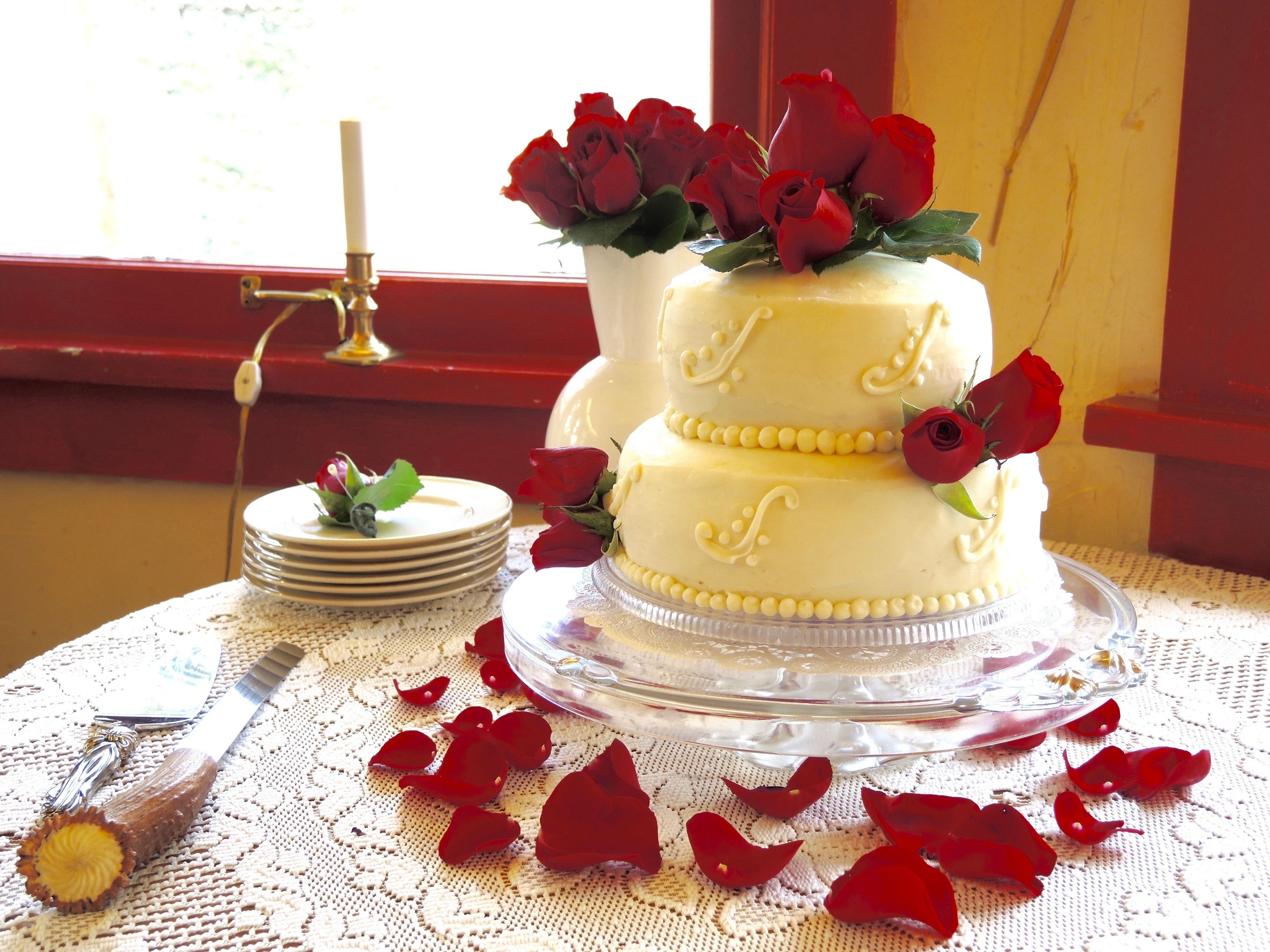 A two-tiered wedding cake with white frosting and red roses on top, placed on a lace tablecloth.