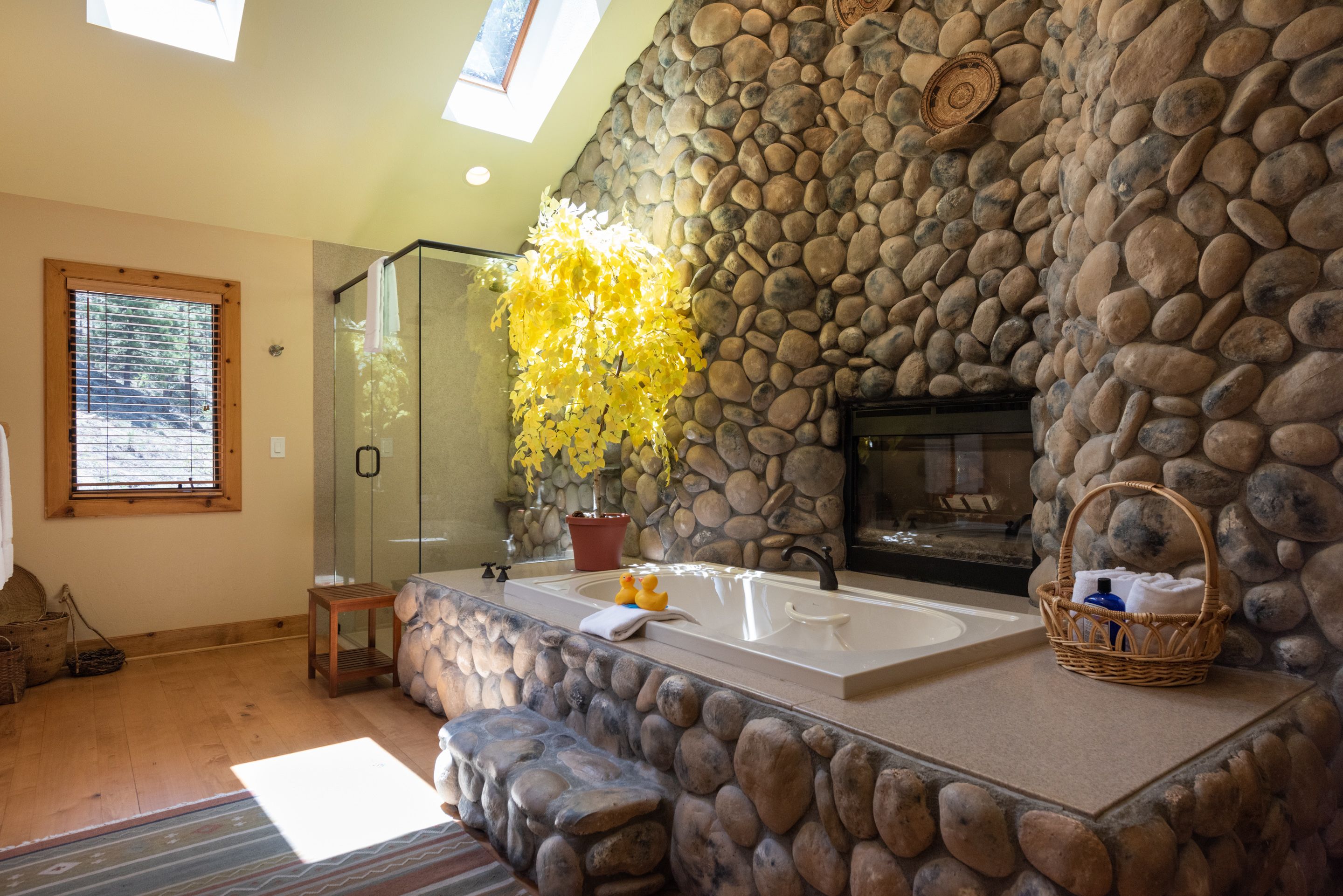 Une salle de bain avec un mur en pierre, une grande fenêtre, une douche et un lavabo agrémenté d’une plante jaune.