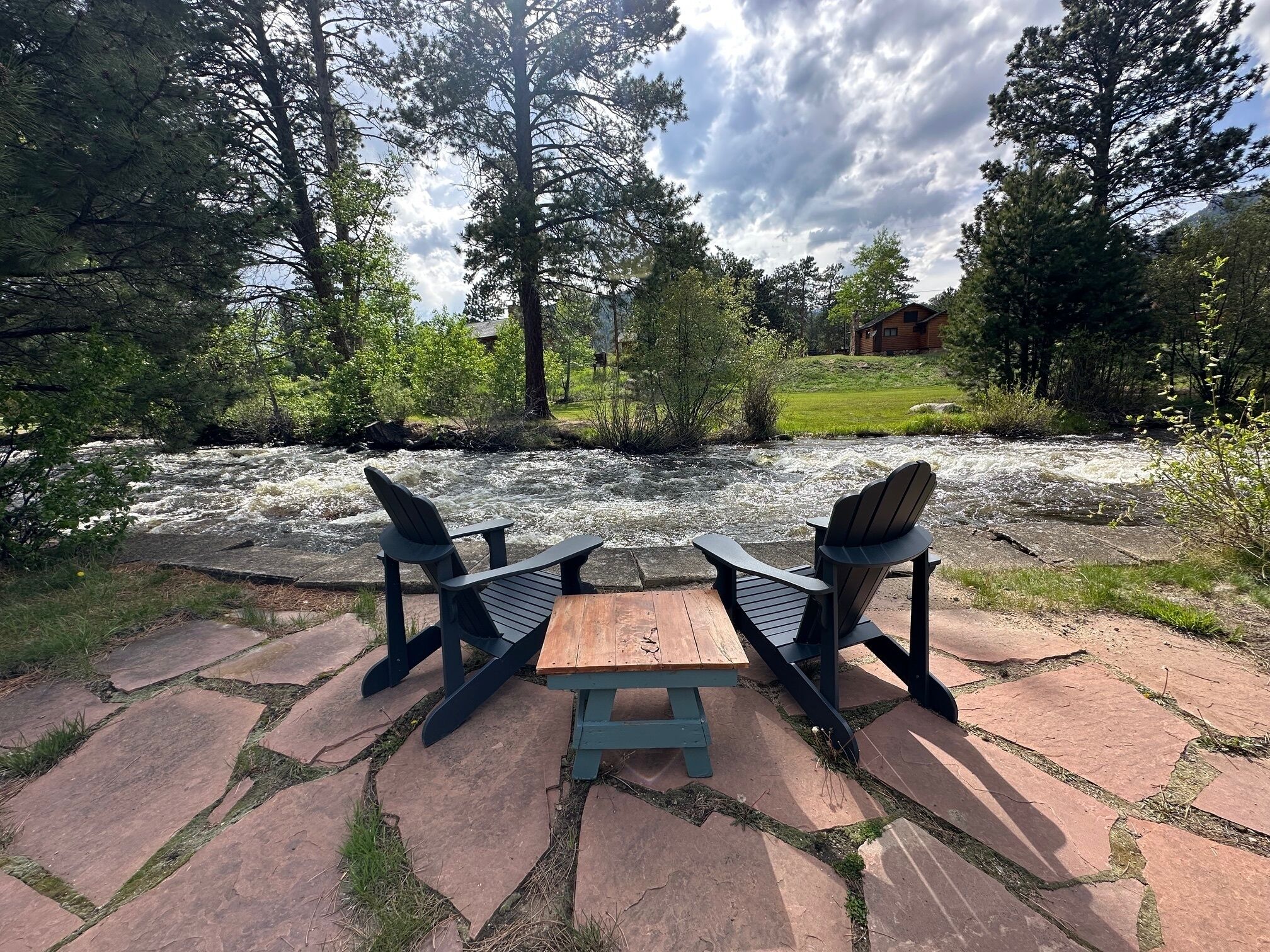 Zona relax lungo il fiume con poltrone Adirondack e un tavolo di legno, circondata da lastricato in pietra e vegetazione.