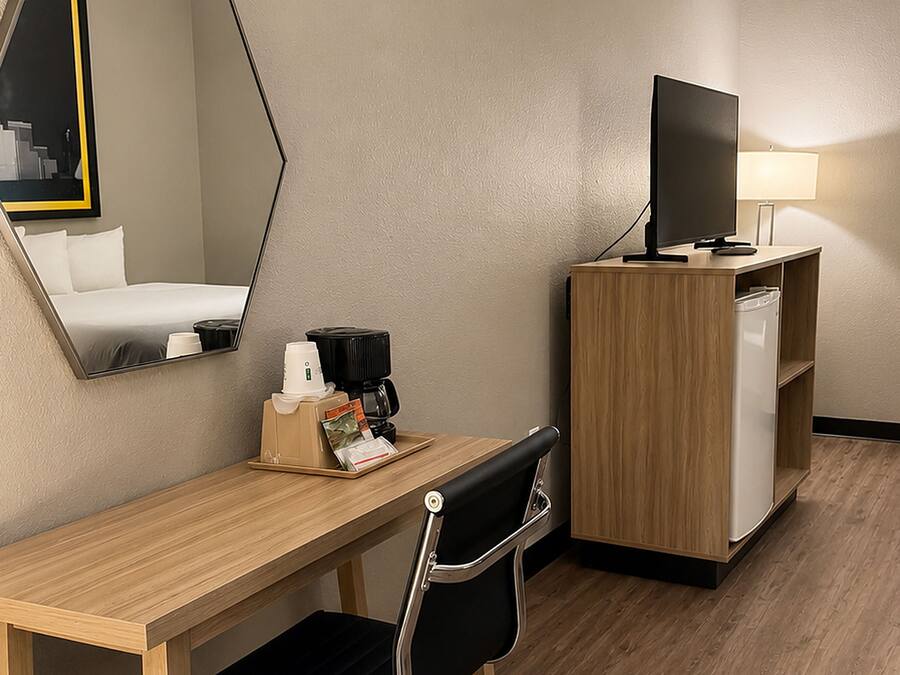 Habitación de hotel con escritorio de madera, una silla, cafetera, televisor sobre un mueble de madera y un espejo en la pared.