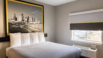 Una habitación de hotel con una cama, un cuadro de un paisaje urbano, una ventana con vistas y un aire acondicionado de pared.