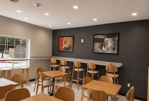 Un área de comedor con mesas y sillas de madera, una ventana con vistas, y dos obras de arte enmarcadas en la pared.