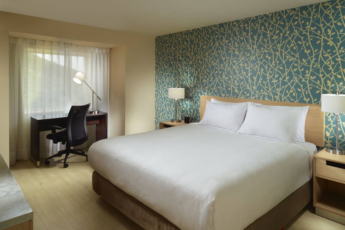 A hotel room with a large bed, a desk, a chair, a nightstand, and a lamp.