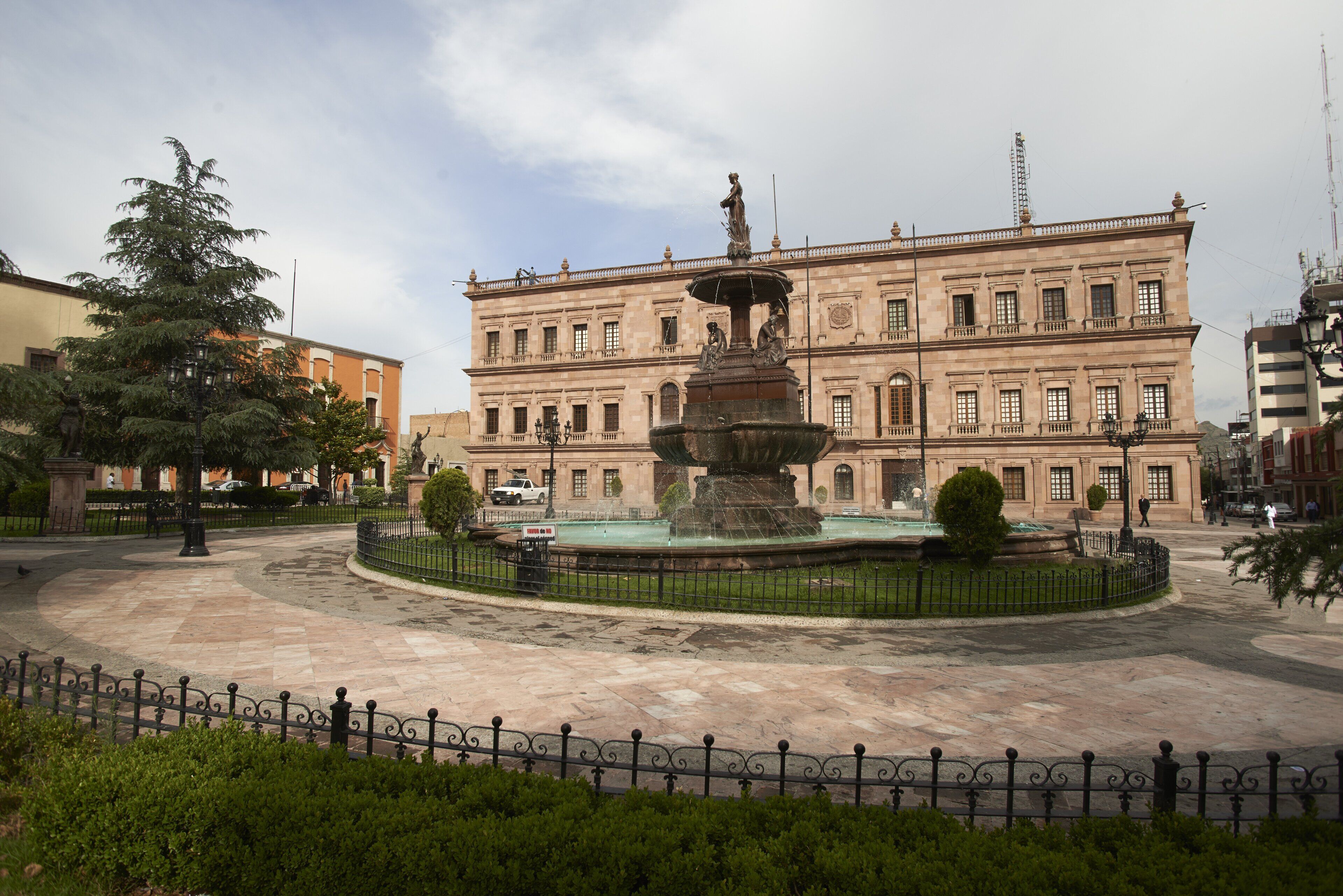 A historic building with a central fountain, surrounded by a paved courtyard and a decorative fence.