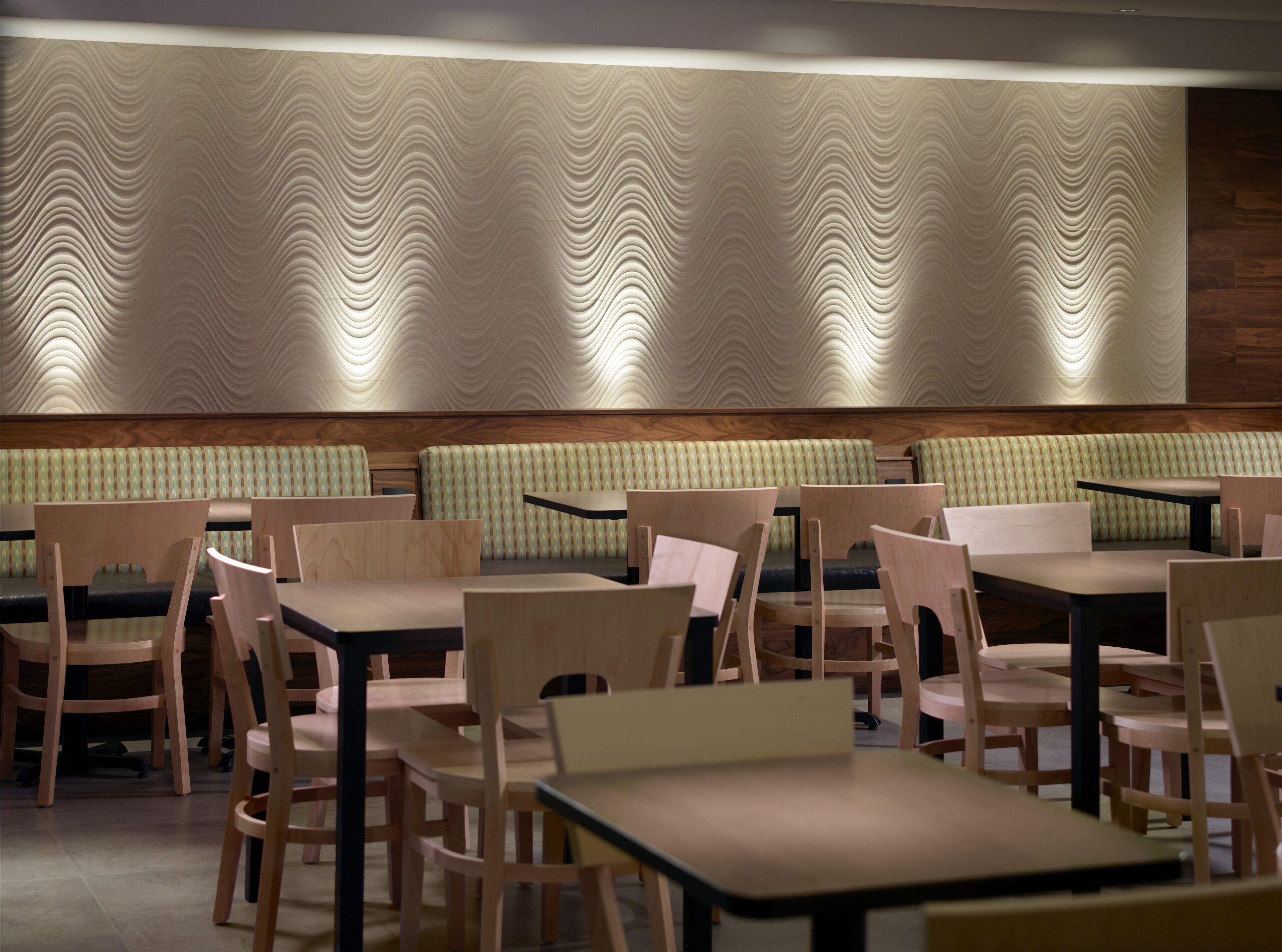 Interior de restaurante con mesas y sillas de madera, pared con diseño y una zona de asientos con banco acolchado verde.
