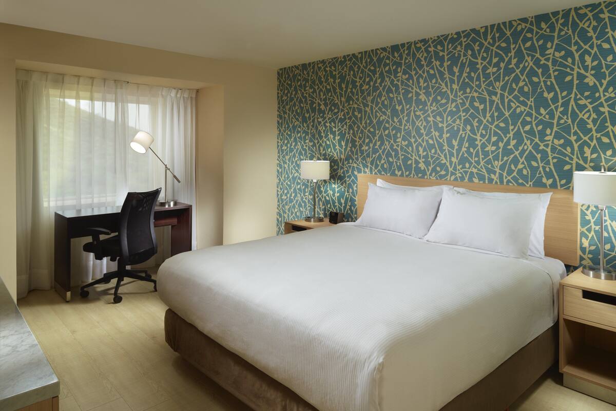 A hotel room with a large bed, a desk with a chair, a nightstand with a lamp, and a window with sheer curtains.