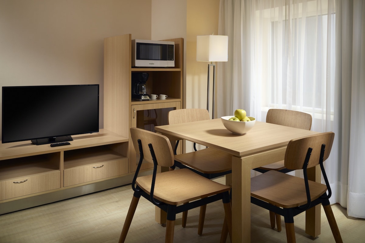 Quarto de hotel com televisão, mesa de jantar com cadeiras, micro-ondas, luminária e janela com cortinas.