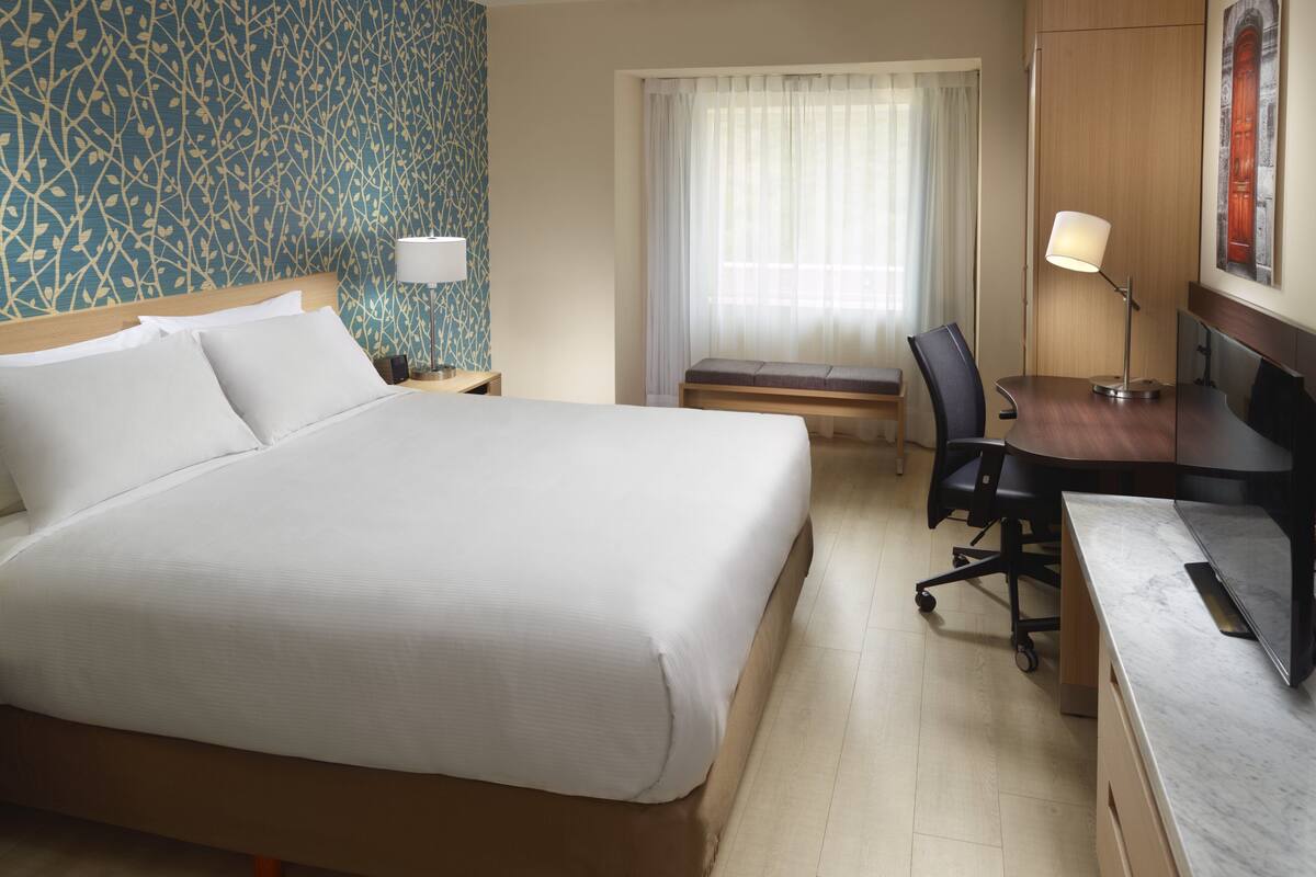 A hotel room with a large bed, a desk with a chair, a lamp, a bench, and a window with curtains.