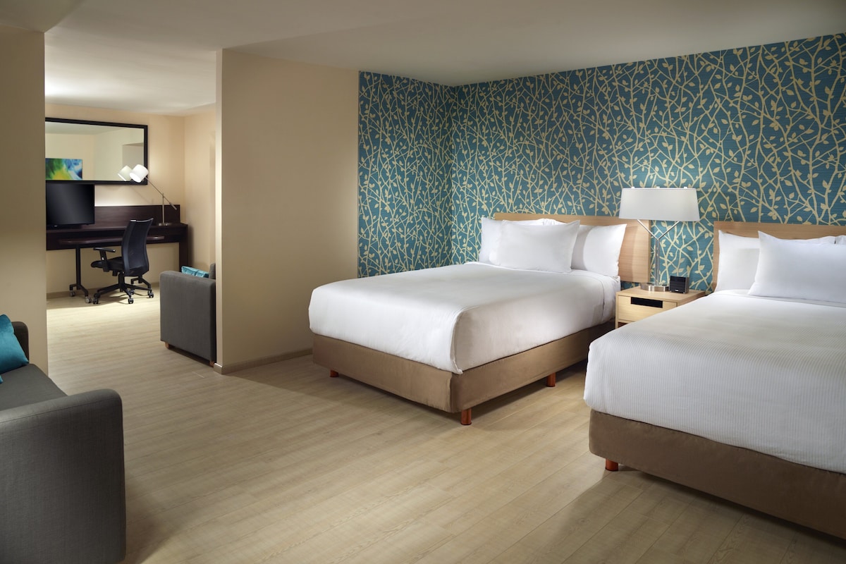 Quarto de hotel com duas camas, uma escrivaninha com cadeira, um espaço de estar pequeno e parede com padrão.