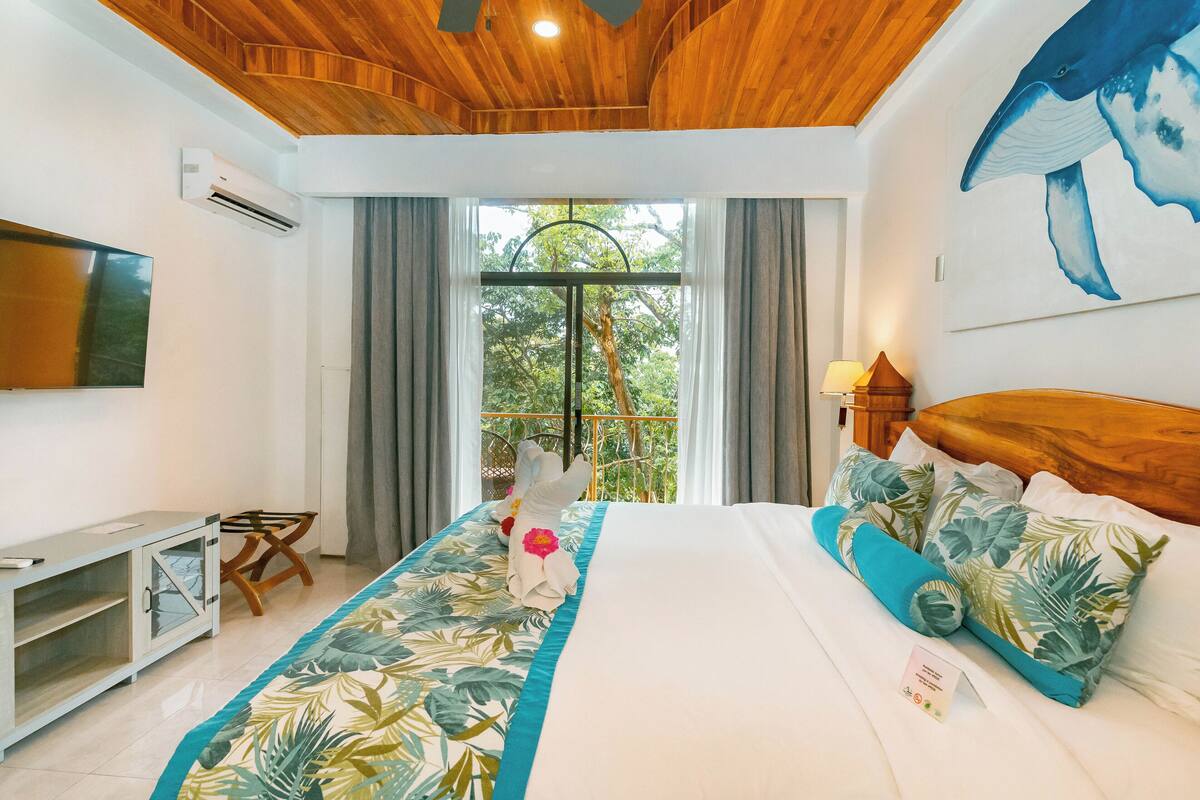 A hotel room with a wooden ceiling, a bed with tropical-themed bedding, a TV on the wall, and a window with curtains.