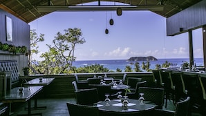 Un espace de restauration en plein air, avec des tables et des chaises, offrant une vue panoramique sur l’océan, avec des palmiers et une île au loin.