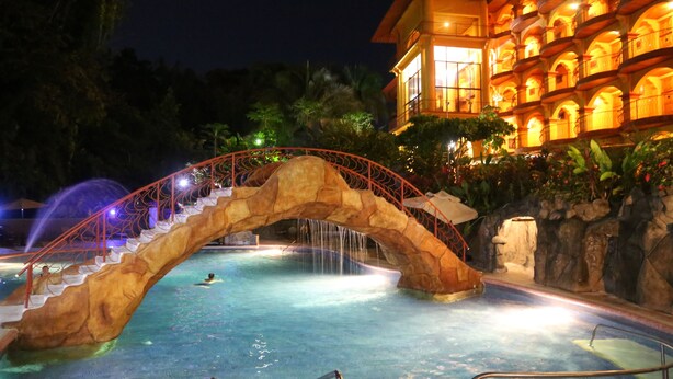 Une piscine avec un pont en pierre, entourée d’un bâtiment doté de nombreux balcons et d’un éclairage chaleureux.