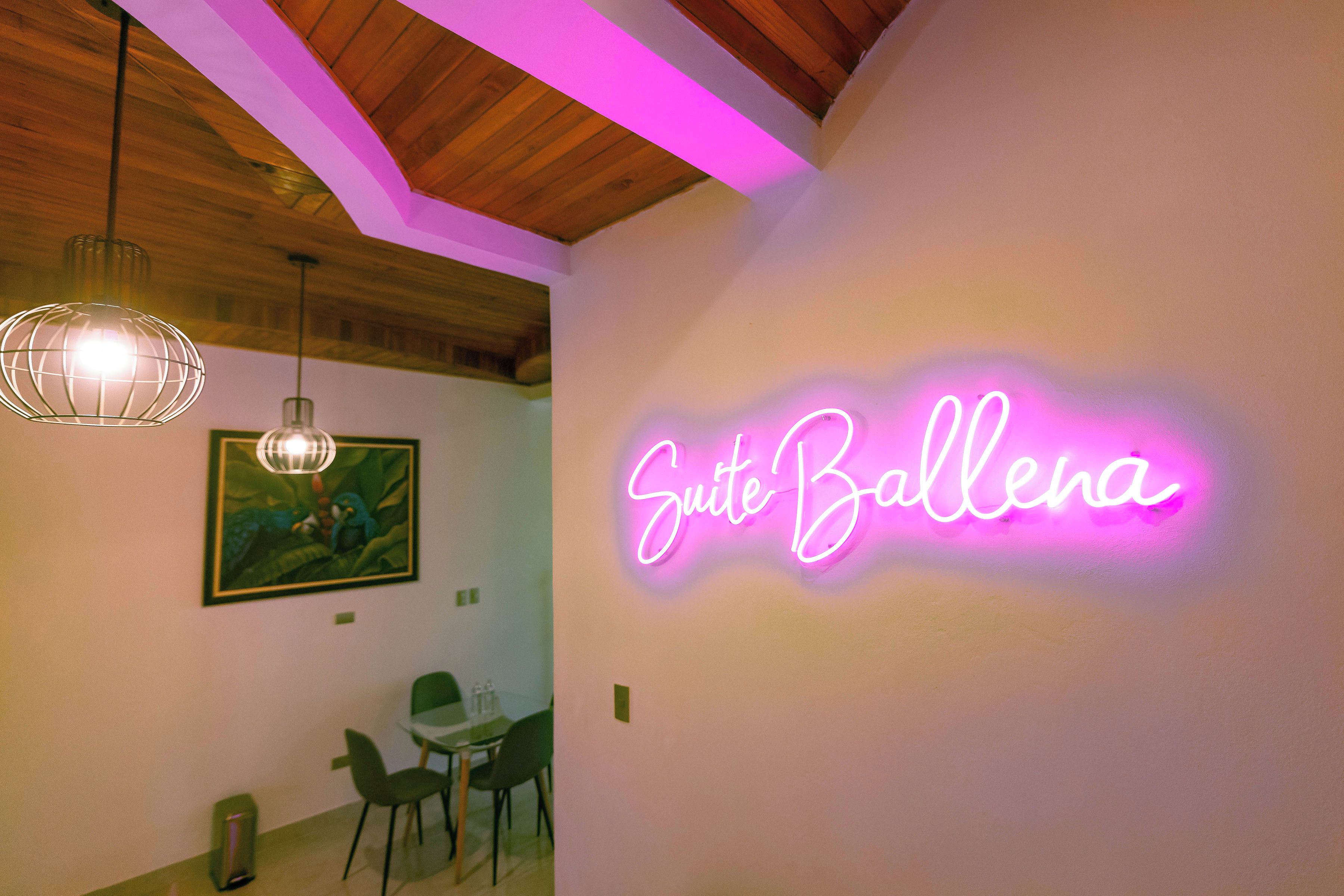 A room with a neon sign reading 'Suite Ballena,' a dining area with chairs and a table, and a hanging light fixture.