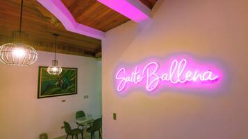 A room with a neon sign reading 'Suite Ballena,' a dining area with chairs and a table, and a hanging light fixture.
