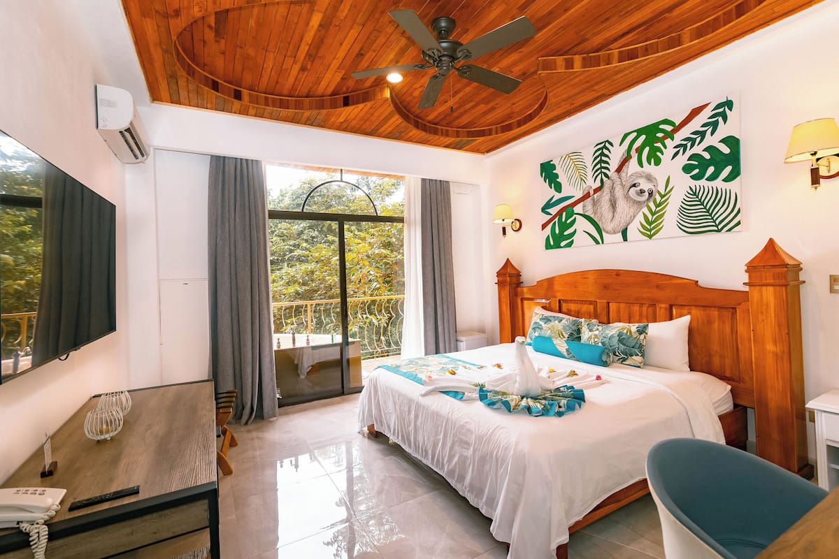 A hotel room with a wooden ceiling, a bed with a tropical-themed headboard, a balcony with curtains, and a large window.