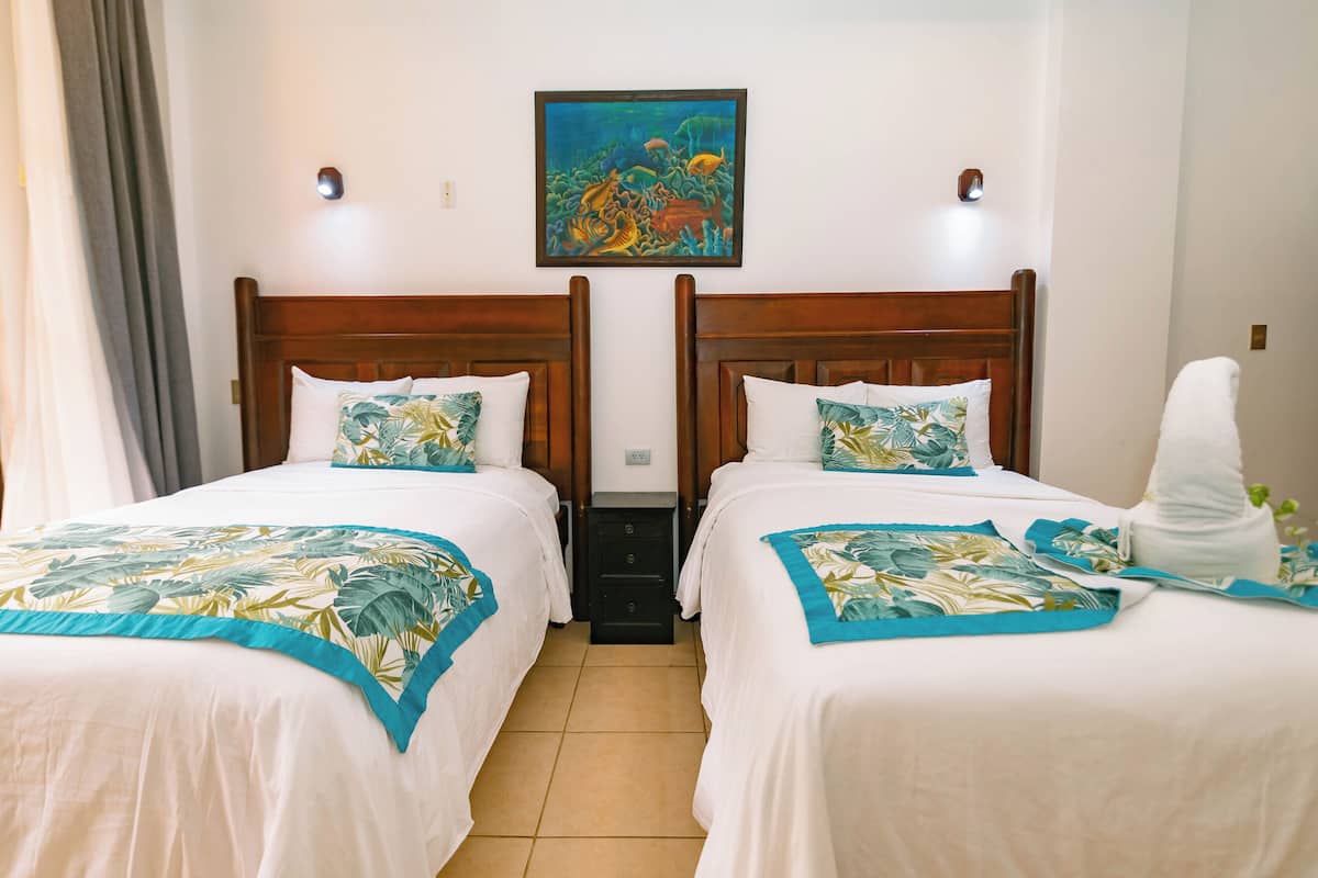 A hotel room with two beds, each with a wooden headboard, white bedding, and a tropical leaf-patterned throw. A painting of a coral reef hangs above the beds.