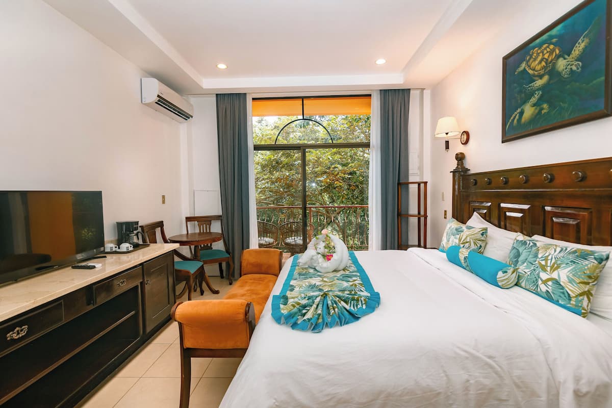 A hotel room with a bed, a TV, a desk, and a window with a view of trees.