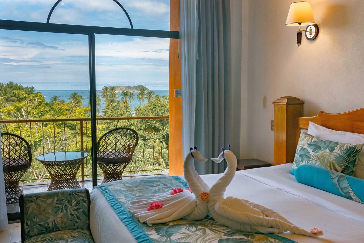 A hotel room with a balcony, a bed with a floral bedspread, two swan-shaped towels, a wooden headboard, and a view of the ocean and palm trees.