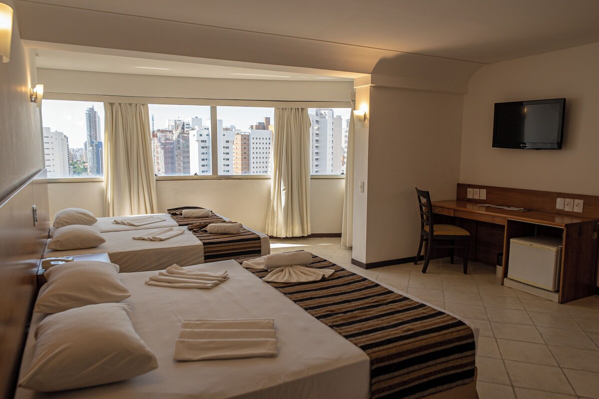 A hotel room with two beds, a desk, a chair, a television, and a view of the cityscape.