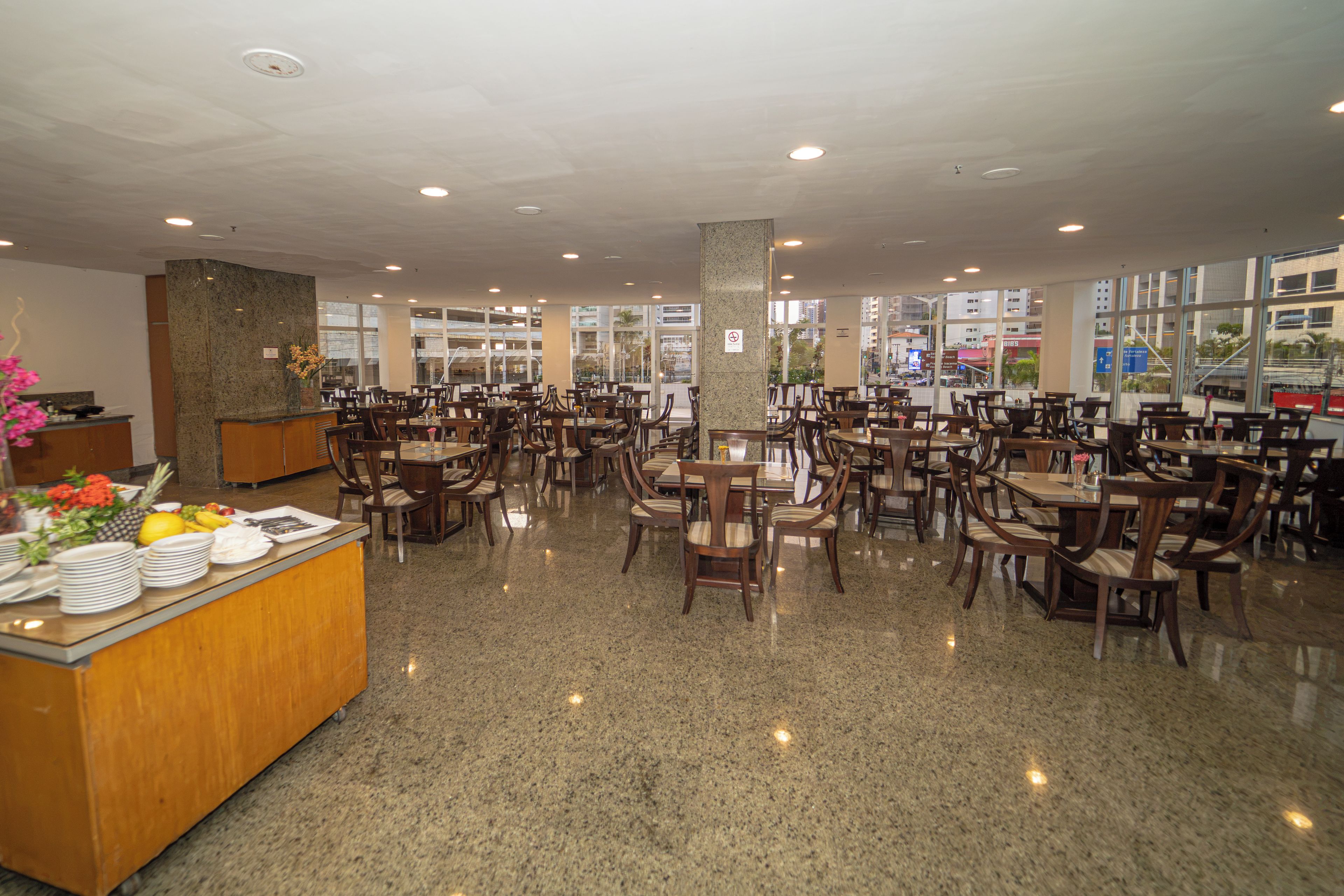 Un restaurante amplio con varias mesas y sillas, una recepción y grandes ventanales con vistas a la ciudad.