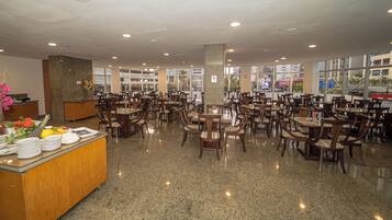 Un restaurante amplio con varias mesas y sillas, una recepción y grandes ventanales con vistas a la ciudad.