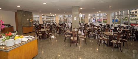 A spacious restaurant with multiple dining tables and chairs, a reception desk, and large windows offering views of the city.