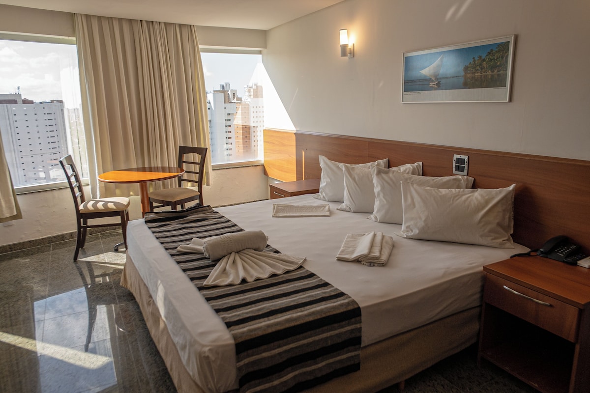 A hotel room with a bed, a chair, a table, a nightstand, a painting, and a view of the city through the window.