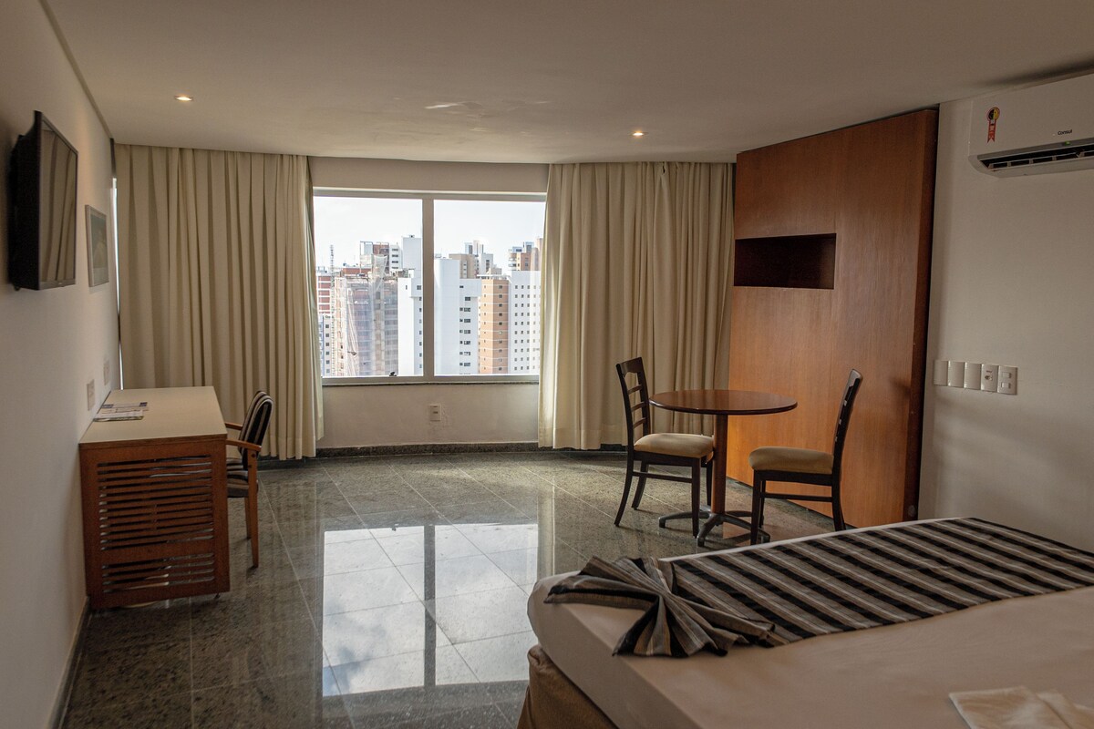 A hotel room with a bed, a desk, a chair, a small table, and a window with curtains.