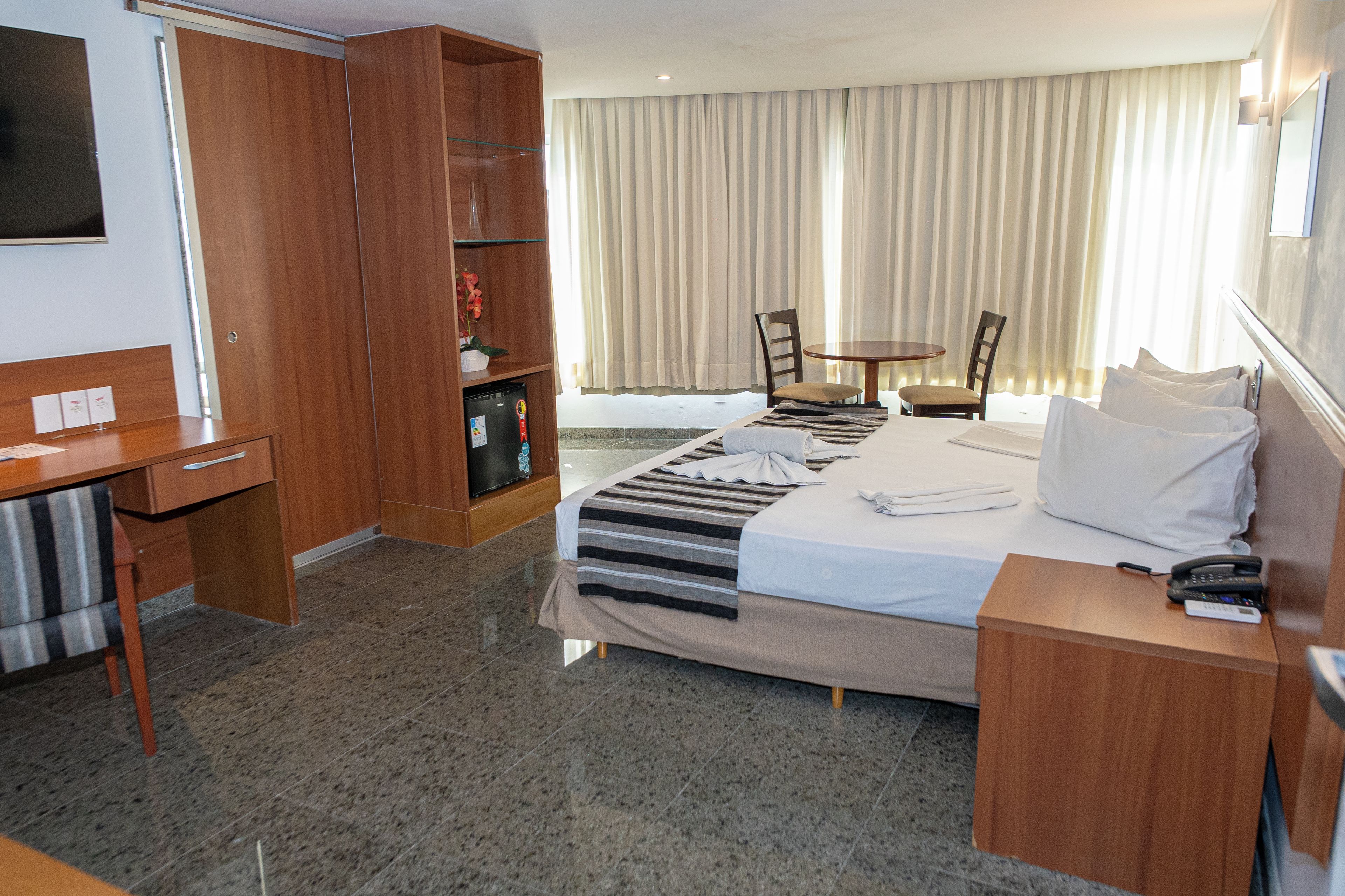 A hotel room with a bed, a desk, a chair, a small table, and a TV.