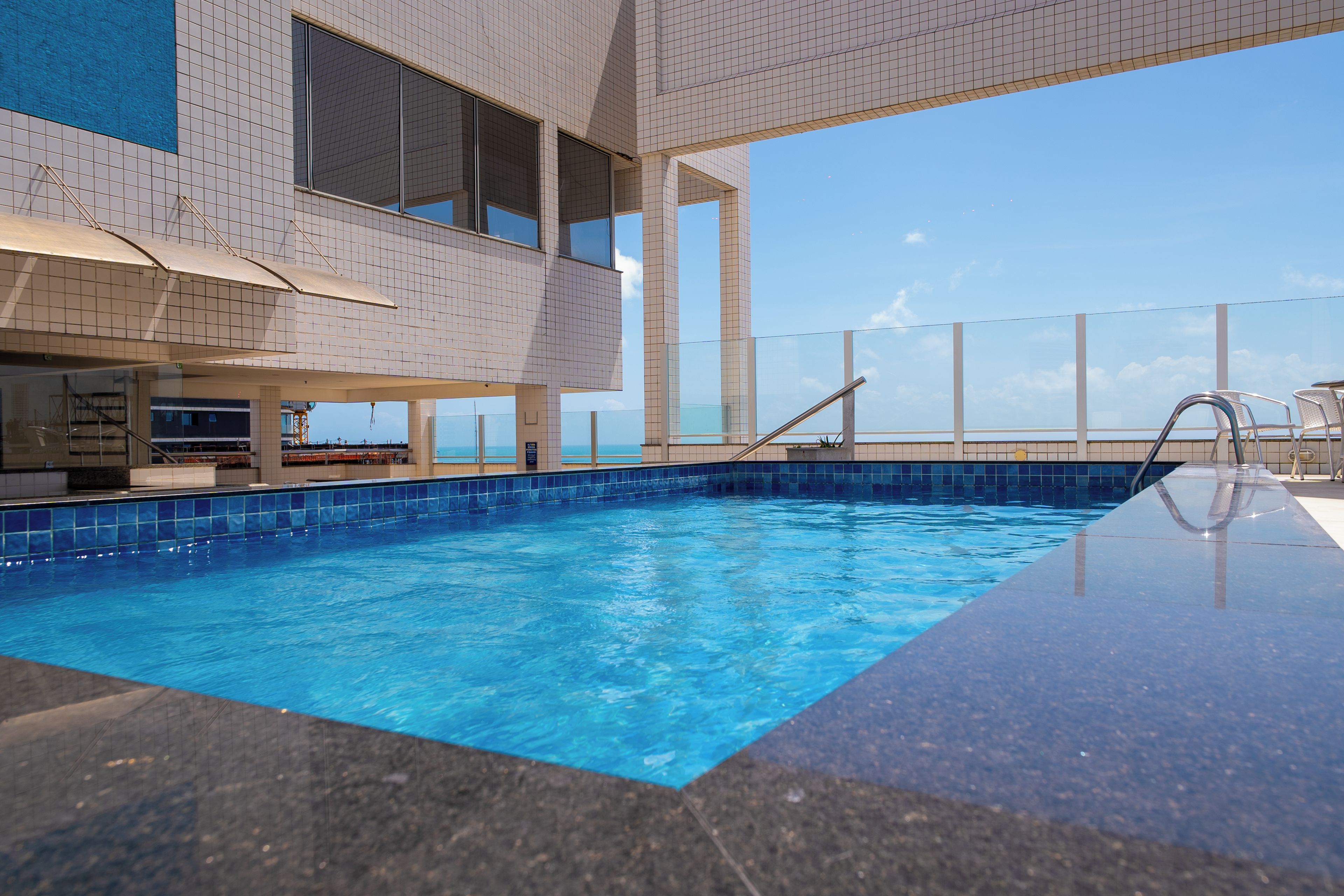 An indoor swimming pool with clear blue water, surrounded by a tiled wall and a balcony with seating.