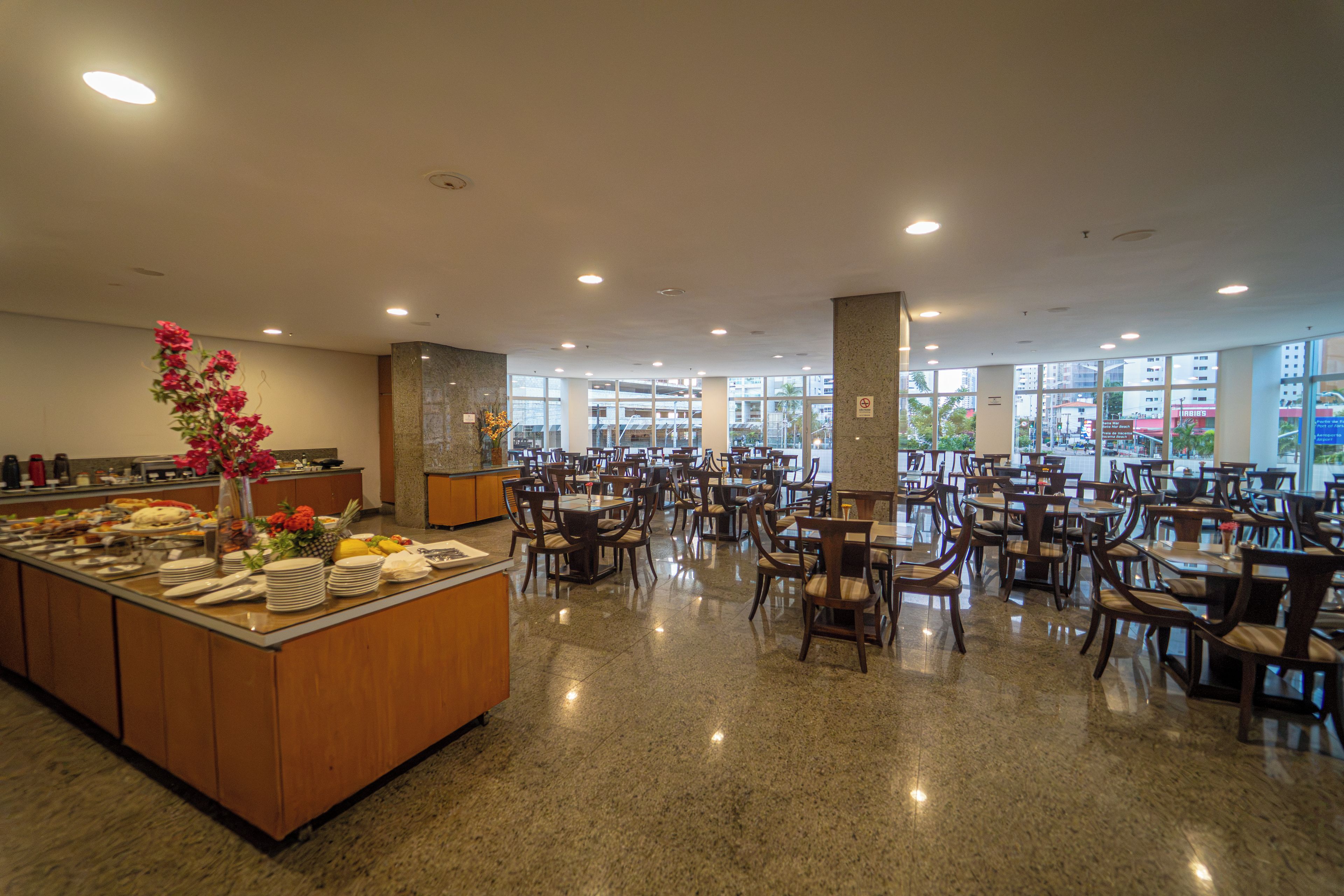 A spacious dining area with multiple tables and chairs, a buffet counter with various food items, and large windows providing a view of the outside.
