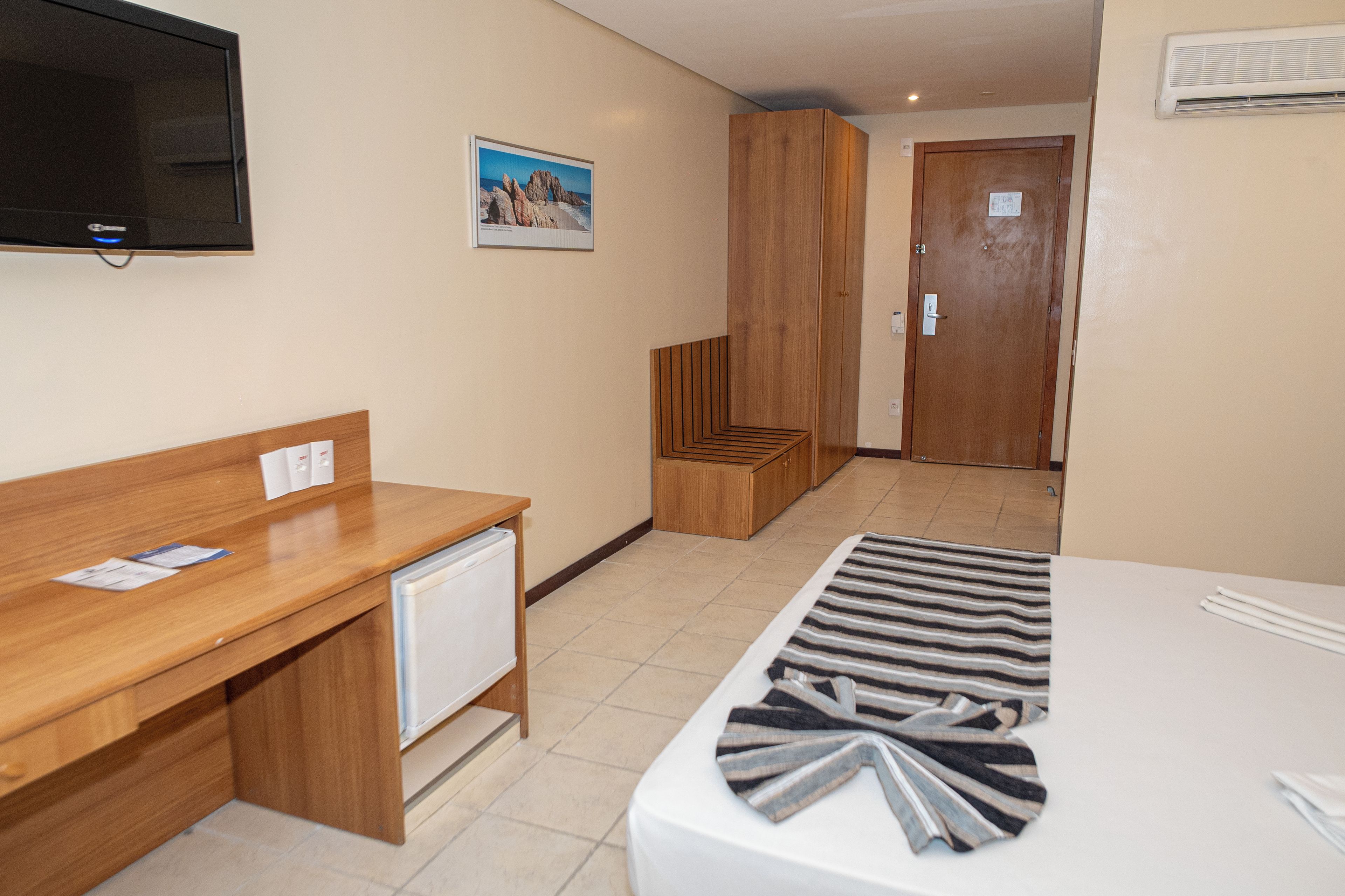 Une chambre d’hôtel avec un bureau en bois, un lit recouvert d’une couverture à rayures, un téléviseur fixé au mur et un tableau encadré au-dessus du bureau.